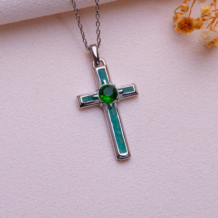 Silver Birthstone Cross Necklace with a green round center stone and green rectangular accents on pale fabric