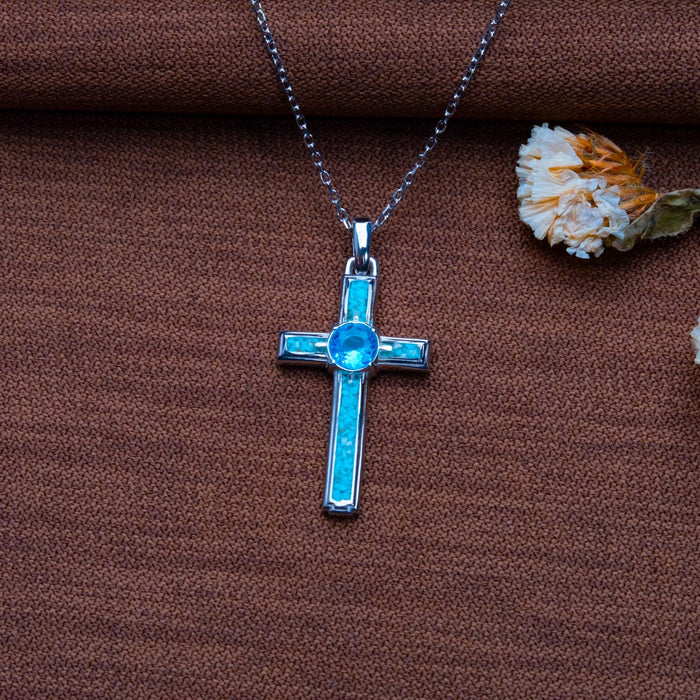 Silver Birthstone Cross Necklace with a bright blue round center stone and blue rectangular accents on brown textured fabric