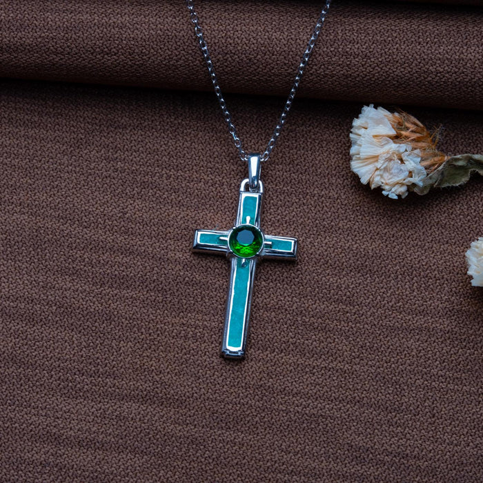 Silver Birthstone Cross Necklace with green round center stone on brown textured fabric beside dried flowers
