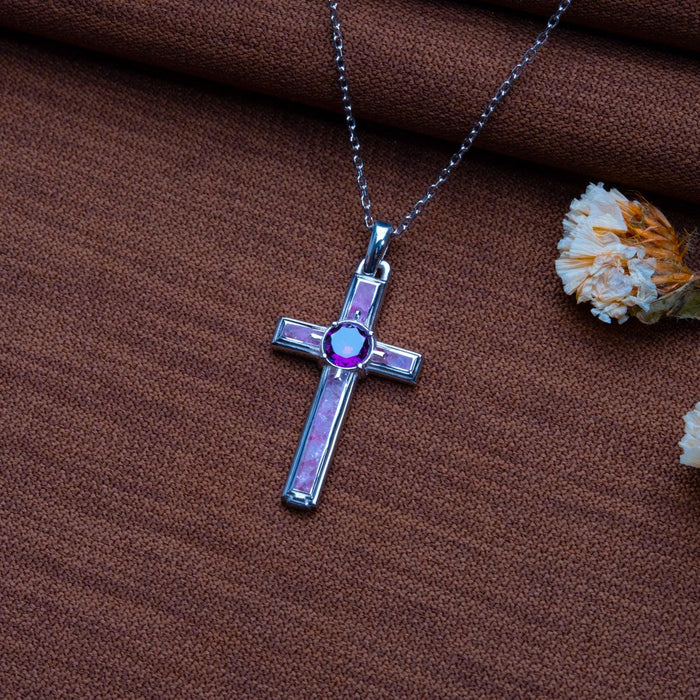 Close view of a dainty amethyst cross necklace with rectangular inset panels on brown textured fabric beside dried flowers