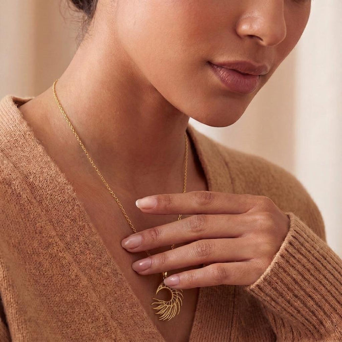 Close cropped model image showing a gold-toned crescent feather pendant necklace with a warm neutral sweater