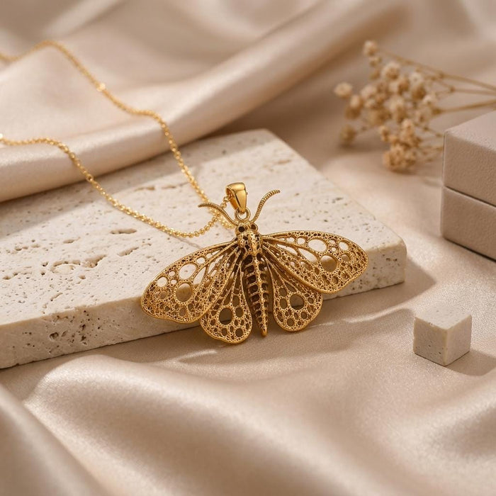 Gold Plated Silver fossil moth necklace with openwork wings displayed on a pale porous stone slab with satin fabric and dried flowers
