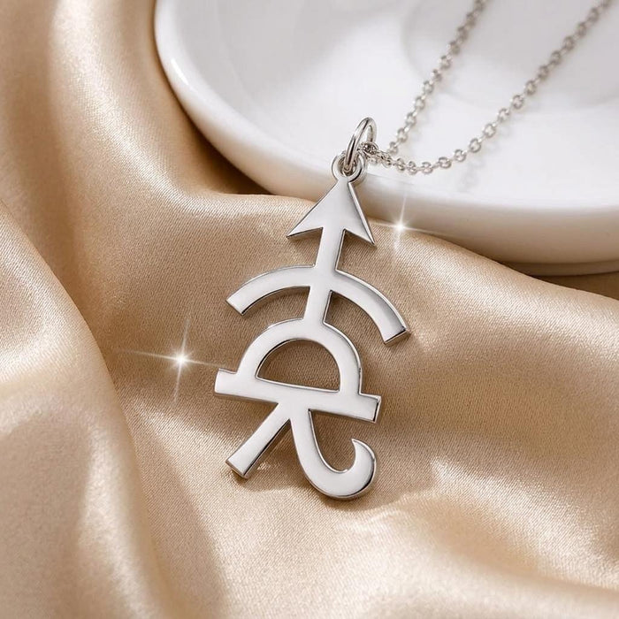 Close-up of the Achilles symbol pendant on champagne satin beside a white dish, showing the crossbar and curled lower terminal