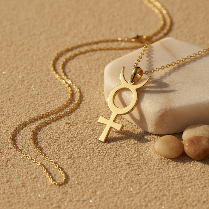 Gold Plated Silver Antimony Symbol Necklace resting on sand beside pale stones