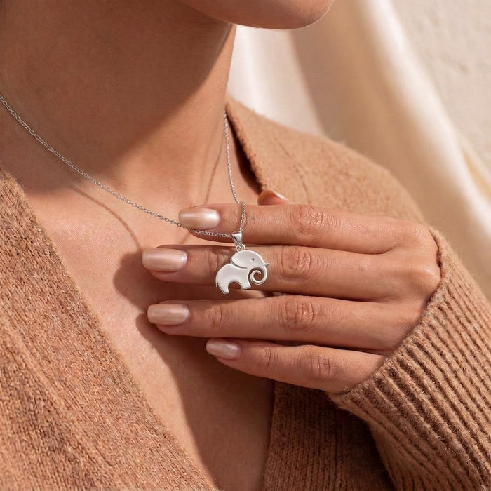Close-up of a solid elephant pendant necklace with an inward-curled trunk held at the neckline over a peach knit cardigan