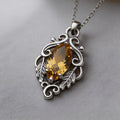 Silver-finish Marquise Yellow Topaz Necklace with a yellow marquise-shaped center stone, visible prongs, scrollwork frame, and leaf details on a gray surface