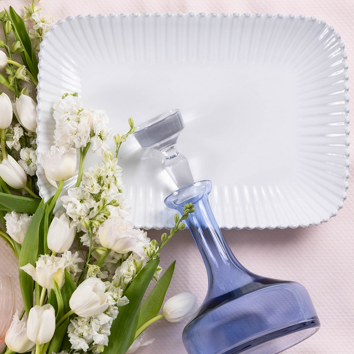 Large rectangular pearlized white serving platter with scalloped edges on pale pink surface with white flowers and blue.