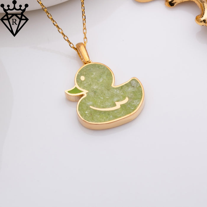 Styled view of a gold-tone duck pendant necklace with pale green textured detail and decorative gold-tone props