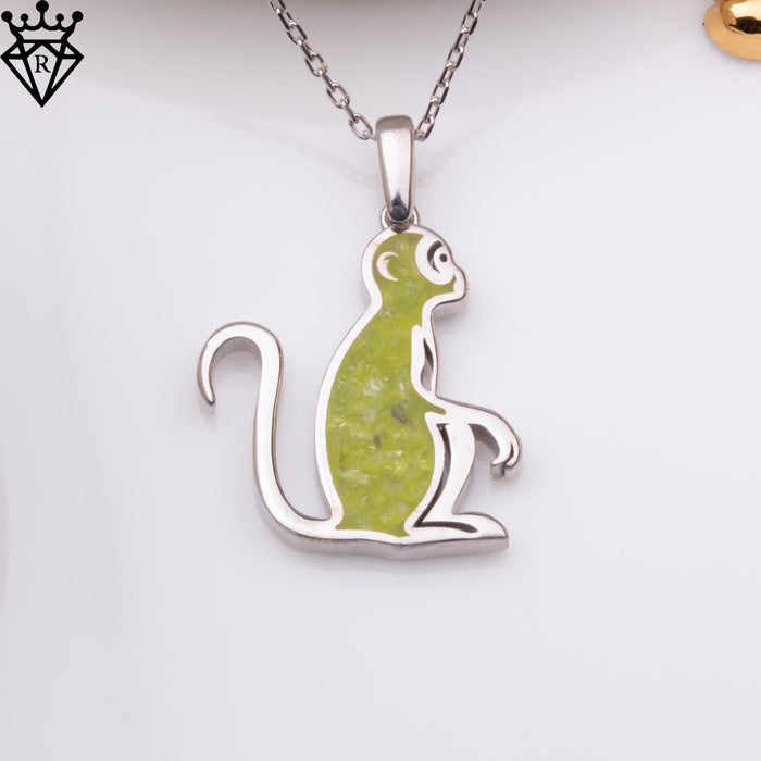 Front product close-up of a seated monkey pendant with a green center detail on a silver chain. A black crown-and-diamond logo watermark appears in the upper-left corner