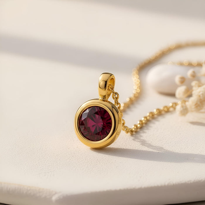 Gold Plated Silver Birthstone Pendant Necklace with a round deep red stone on a pale textured surface