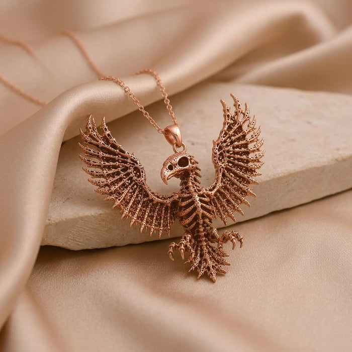 Rose-toned Sacred Eagle Skeleton pendant displayed on a pale textured stone slab with draped champagne satin
