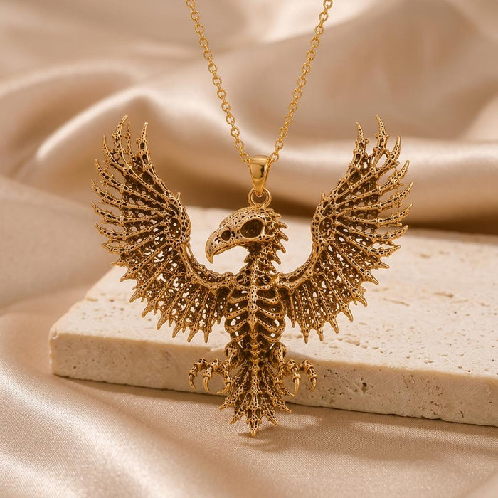Front-facing gold-toned Sacred Eagle Skeleton pendant on a pale stone surface with champagne satin behind it