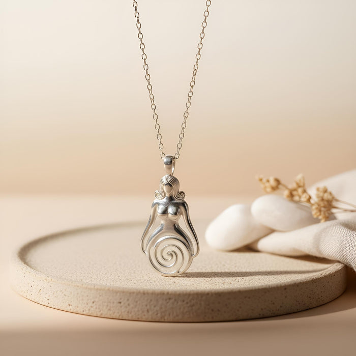 Silver Spiral Goddess Necklace displayed upright on a textured round platform beside white stones and dried stems