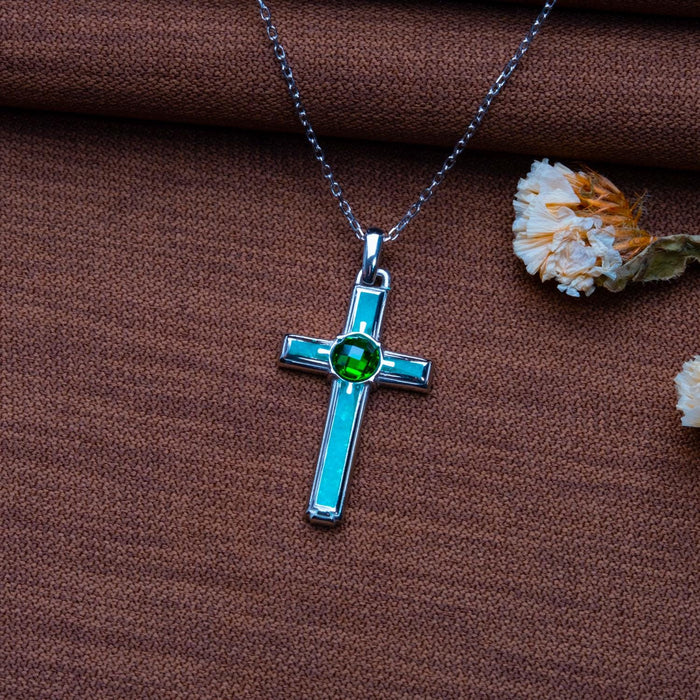 Rectangular aventurine cross necklace with teal inset panels and a round green center stone on brown textured fabric beside dried flowers