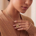 Close neckline view of a model holding a Gold Plated Silver sunflower pendant necklace with a red center detail against a tan knit