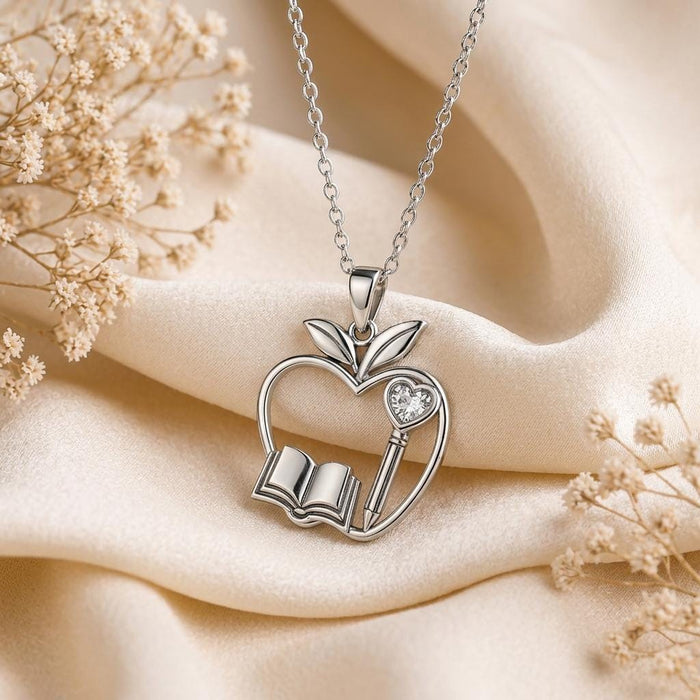 Close-up of the Silver Teacher Apple Necklace with open apple frame, book, pencil, leaves, and clear accent on ivory fabric