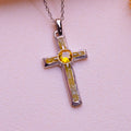 Close-up of the Vertical Framed Birthstone Cross Necklace with framed inset panels and a round yellow center stone on pale fabric
