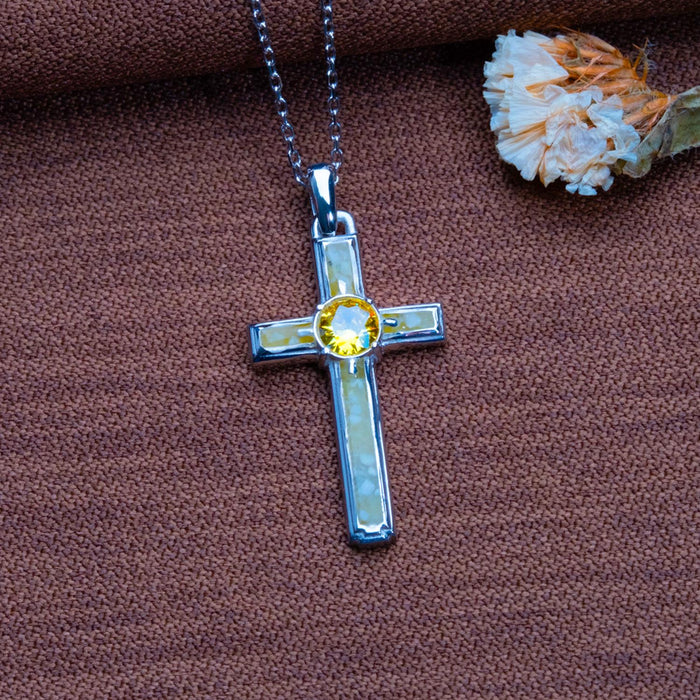 Vertical Framed Birthstone Cross Necklace shown on brown textured fabric beside a dried flower
